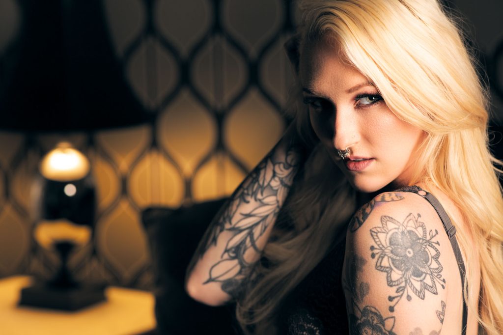 Portrait of Sonoma county boudoir makeup artist Ducky Rickman. She leans back slightly with her head turned toward the camera while wearing a black lace top as warm lighting from a nearby lamp highlights her features against a patterned wallpaper background.