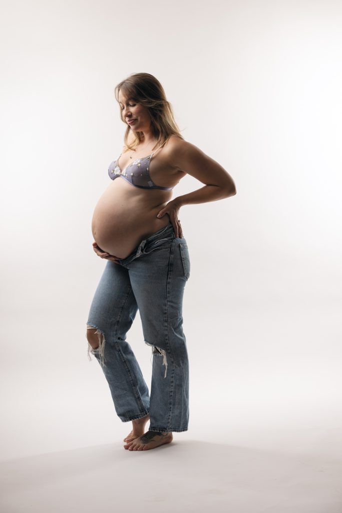 A pregnant woman stands confidently in a bright studio setting for her Sonoma County maternity photos. She turns her body slightly while looking back over her shoulder toward the camera with a gentle smile. One hand rests on her hip as she highlights her rounded belly in a relaxed pose. Soft even lighting illuminates the natural colors of her light purple floral lace bra and blue ripped denim jeans. She remains barefoot against the seamless white backdrop.