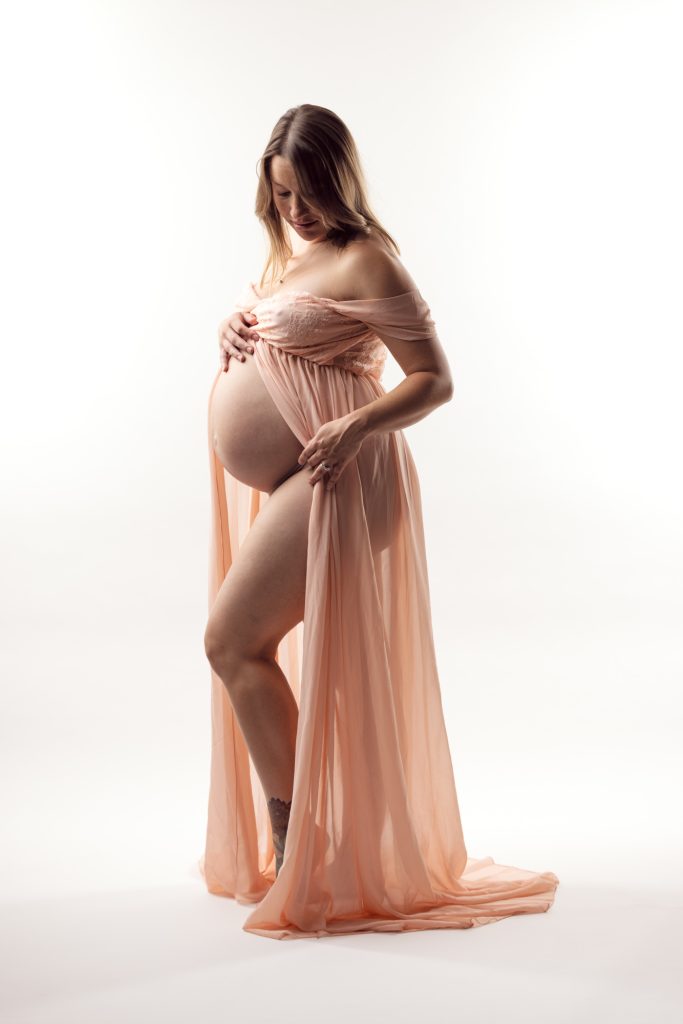 A pregnant woman stands gracefully in a bright studio setting during her Sonoma maternity boudoir photo shoot. She looks down gently while cradling her rounded belly with both hands in a tender pose. Soft even lighting highlights the delicate peach tones and flowing sheer fabric of her off-the-shoulder dress which drapes elegantly around her form with the open front creating beautiful movement. She remains barefoot against the seamless white backdrop.918msFast