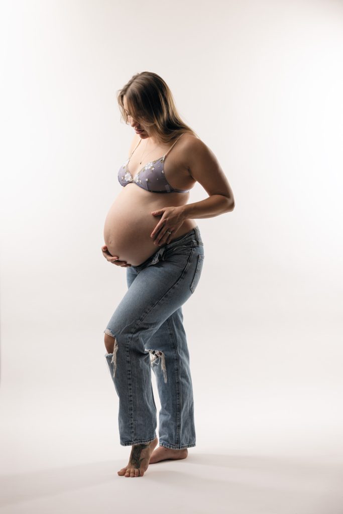 A pregnant woman stands in profile within a bright minimalist studio during her tasteful maternity boudoir in bra and jeans. She looks down gently while cradling her rounded belly with both hands in a protective and relaxed pose. Soft even lighting highlights the natural colors and textures across the image including her light purple floral lace bra and blue ripped denim jeans. She remains barefoot with a visible tattoo on her foot against the seamless white backdrop.