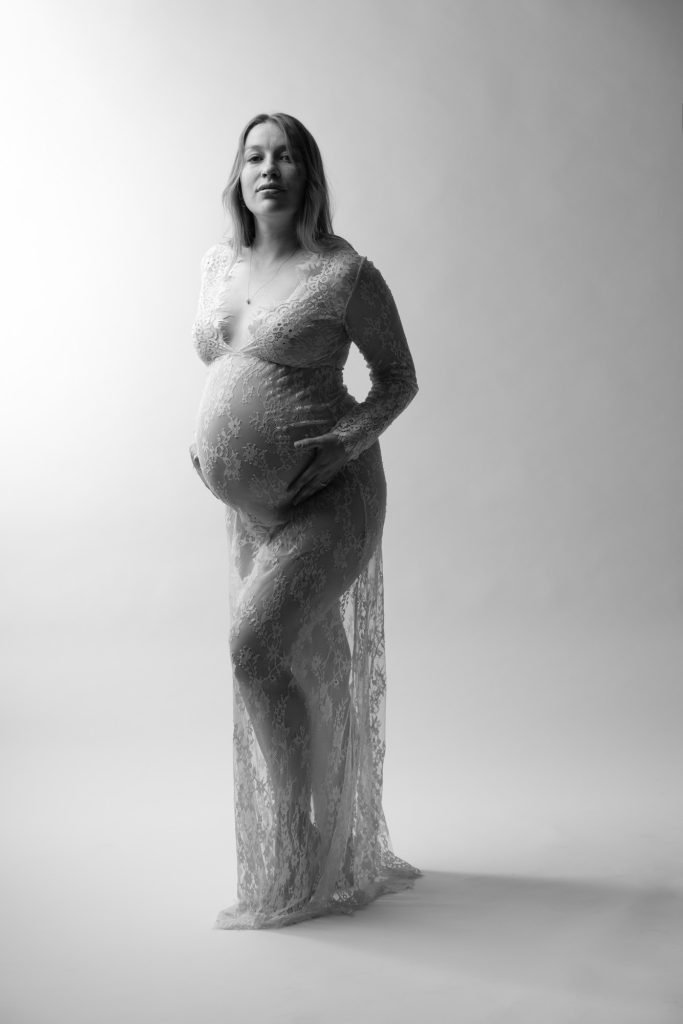 A pregnant woman stands gracefully in a bright studio setting for her Vogue inspired maternity session. She poses with one hand resting gently on her rounded belly while looking directly toward the camera with a poised expression. Dramatic lighting creates strong highlights and shadows across the black and white image emphasizing the intricate lace patterns of her long fitted dress. The sheer fabric drapes elegantly around her form. She remains barefoot against the seamless white backdrop.