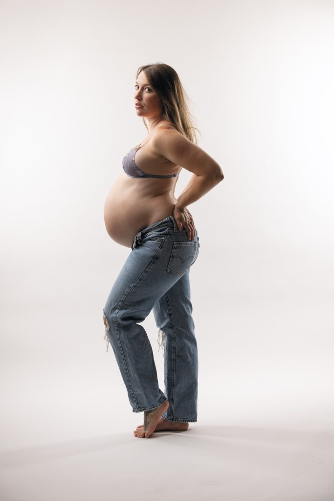 A pregnant woman stands confidently in a bright studio setting during her vogue maternity photo shoot. She turns her body slightly while looking back over her shoulder toward the camera with a direct gaze. One hand rests on her hip as she highlights her rounded belly in a relaxed pose. Soft even lighting illuminates the natural colors of her light purple floral lace bra and blue ripped denim jeans. She remains barefoot against the seamless white backdrop.