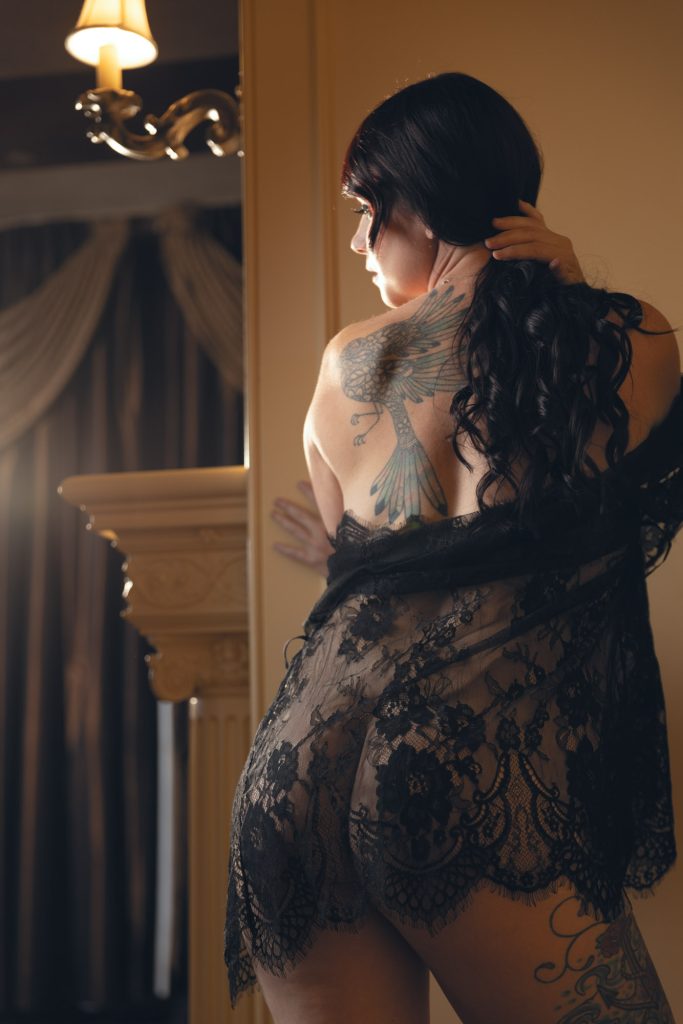 Artistic back pose during a Sonoma County boudoir session featuring a woman with dark wavy hair touches her neck, sheer black lace robe drapes off one shoulder revealing a detailed blue bird tattoo on her back, warm yellow lighting from overhead sconce casts soft glow on ornate pillar, neutral wall, and dark curtains in elegant composition.
